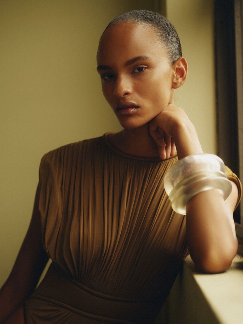Portrait of a Black woman with tan dress and a bracelet posing indoors with natural light, showcasing beauty and fashion.