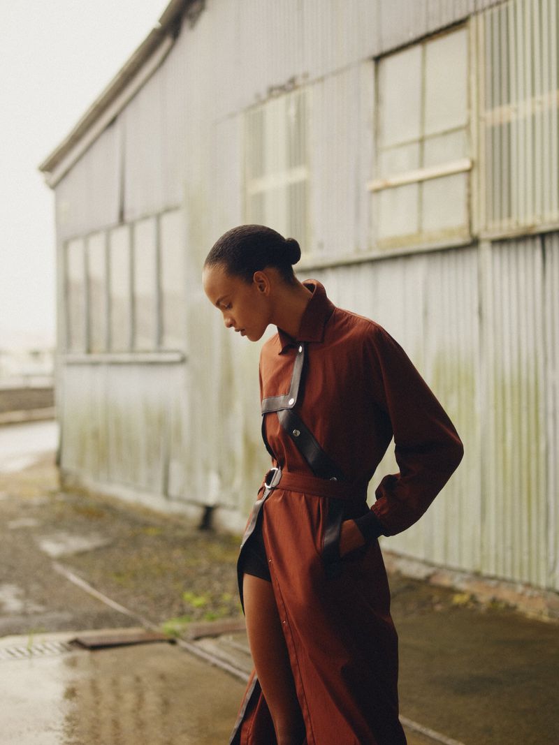 Woman in long brown coat with harness and belt poses in front of weathered metal building, fashion editorial, outdoor shot.