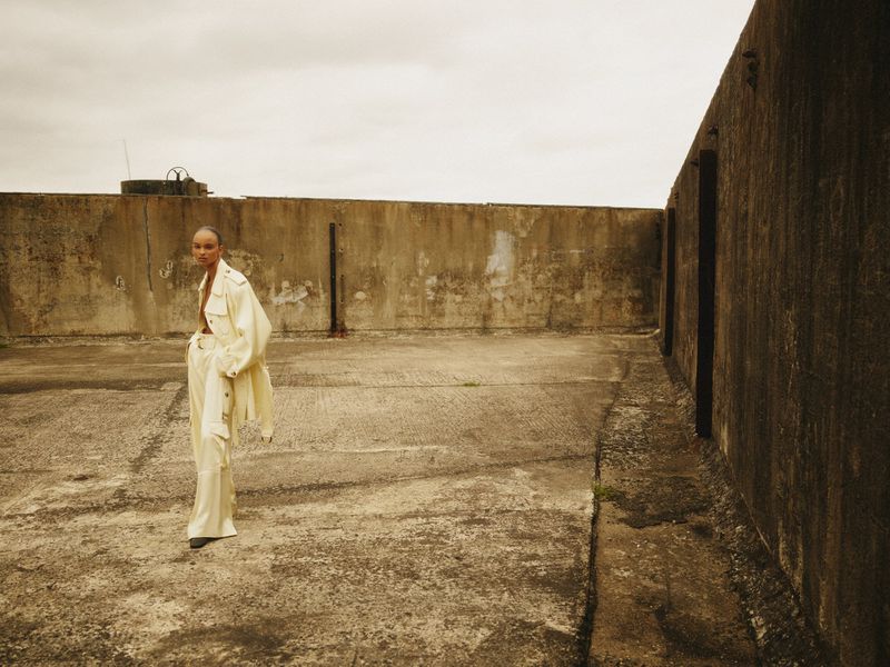 Fashion editorial portrait of an adult female model in a light suit posing on a concrete rooftop with a cloudy sky backdrop.