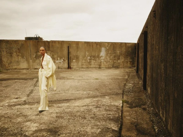 Fashion editorial portrait of an adult female model in a light suit posing on a concrete rooftop with a cloudy sky backdrop.