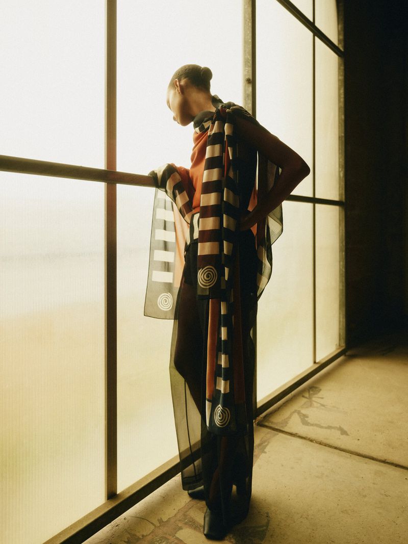 Fashion shot of an adult female standing in front of a window wearing a sheer outfit with graphic patterns in a minimalist interior.
