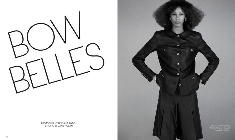 Editorial fashion shot of an adult woman with afro, wearing a black satin suit, posing indoors against a grey backdrop with studio lighting.