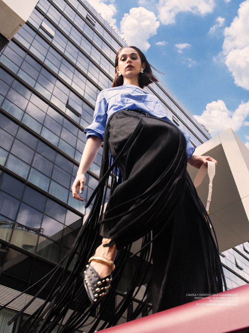 Model in a blue striped shirt and black fringe trousers shot from a low angle against a modern glass office building.