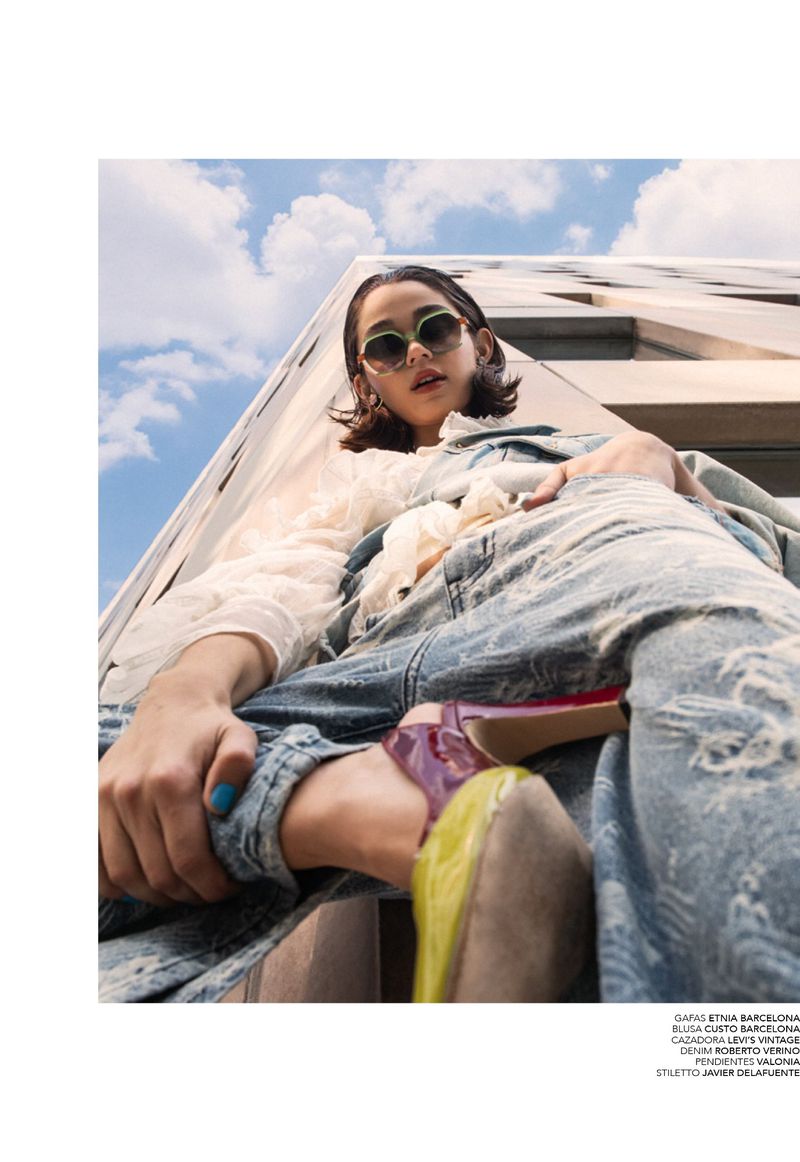 Low-angle shot of a woman wearing sunglasses, a white ruffle blouse, distressed light-blue denim, and colourful heels against a building.