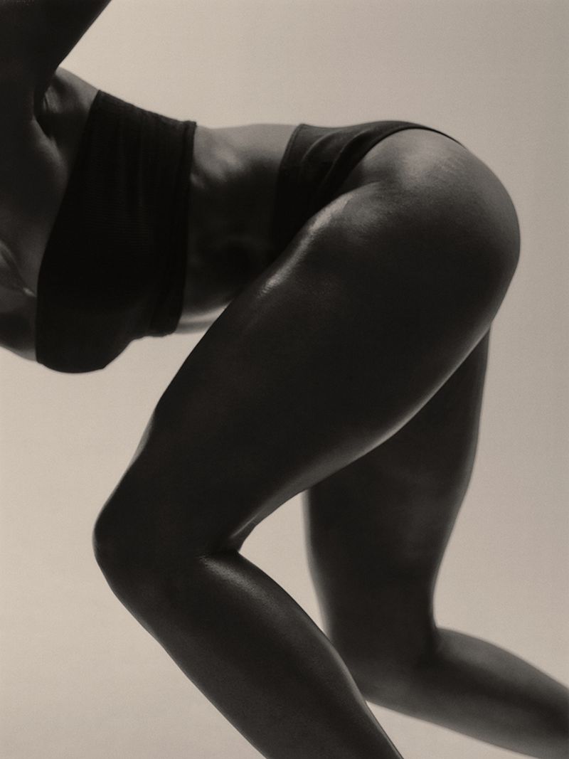 Close-up of a muscular person in a dark athletic top and bottoms posing in a running stance against a plain studio background.
