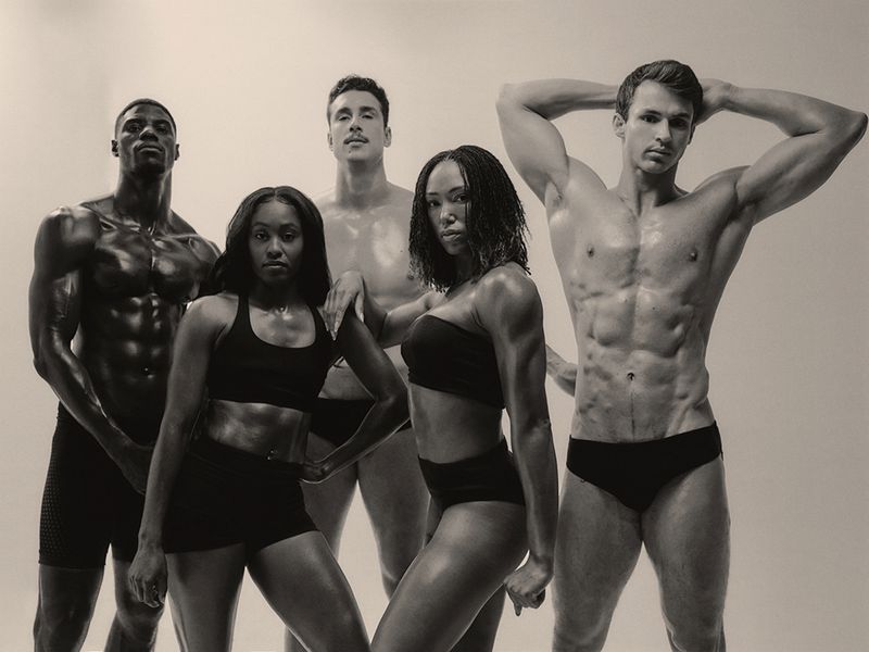 Five muscular athletes posing in black athletic swimwear against a light studio backdrop for a group portrait.