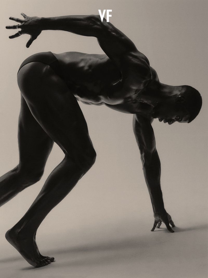 Muscular Black male athlete crouched in a dynamic sprinter start stance against a neutral studio backdrop.