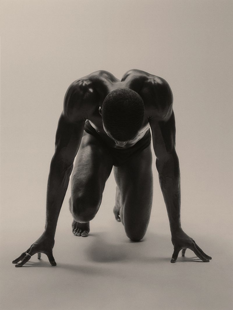 Monochrome image of an athletic African-American man in a sprinter's stance, showcasing musculature in a neutral studio setting.