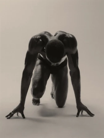 Monochrome image of an athletic African-American man in a sprinter's stance, showcasing musculature in a neutral studio setting.