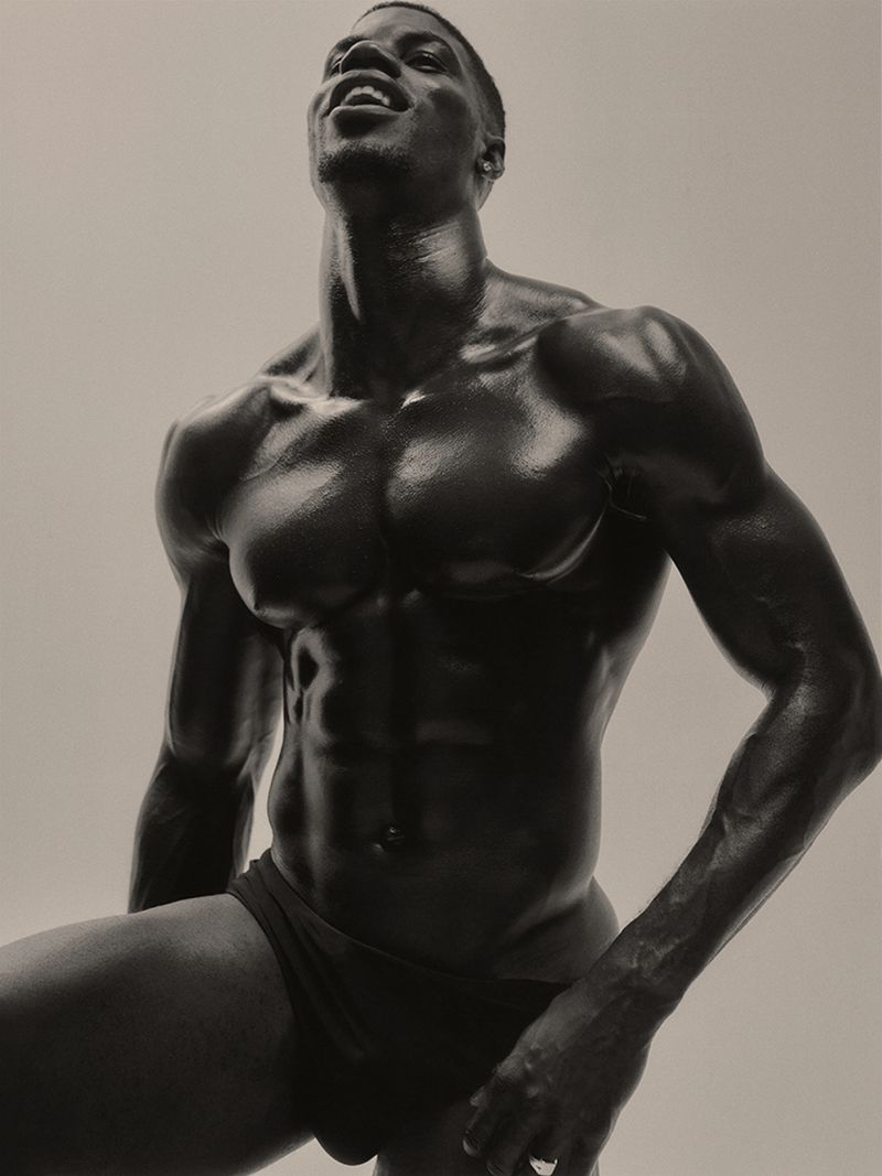 Muscular Black man posing in black briefs with oiled skin against a neutral studio backdrop.