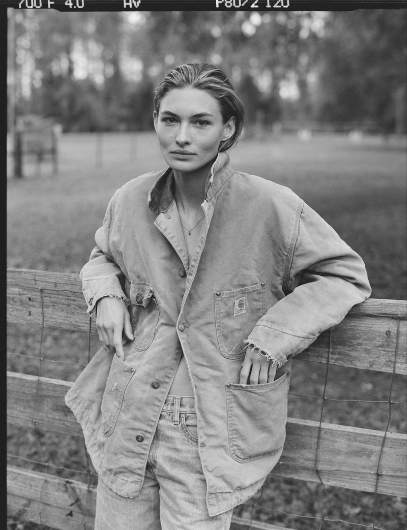 Black-and-white portrait of a woman in an unbuttoned jacket leaning against a wooden fence in a grassy field, looking at the camera.