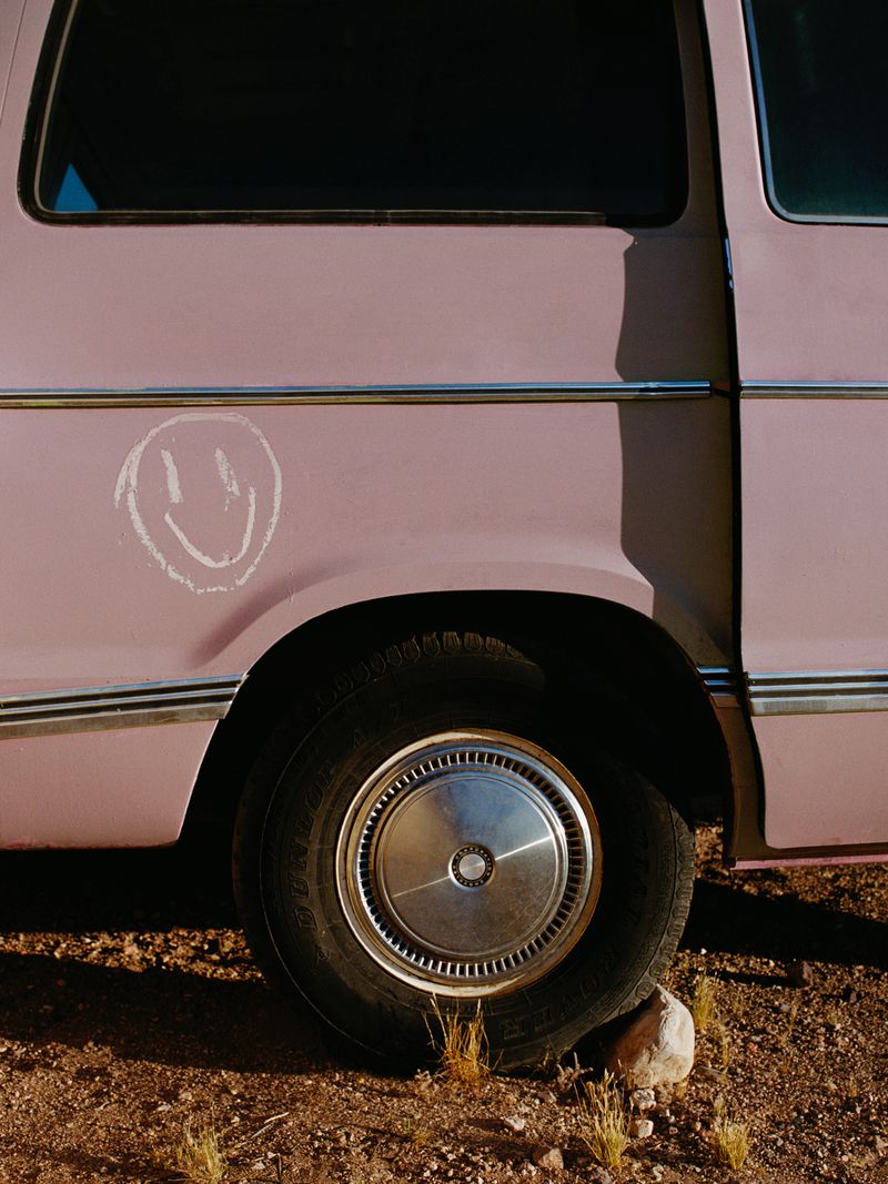 Close-up of a vintage pink van with a drawn smiley face on the body, parked on a dirt road in a desert landscape.