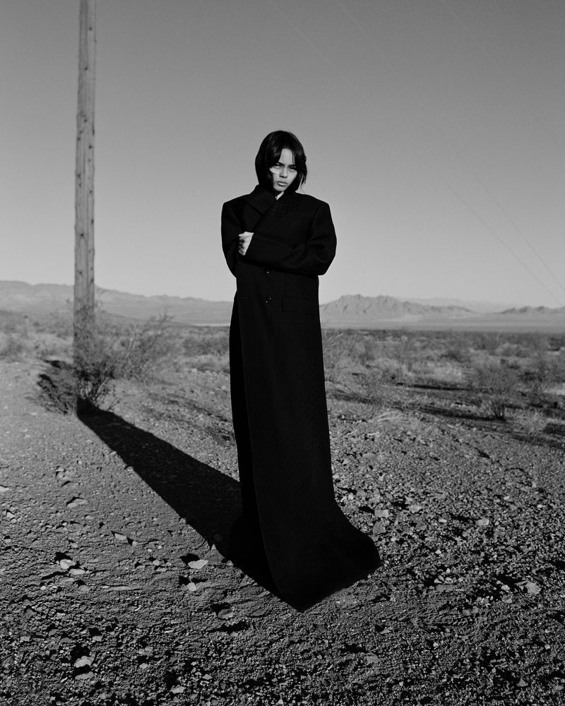 Black and white shot of a woman in a long dark coat standing on a dirt road in a desert landscape with a utility pole nearby.