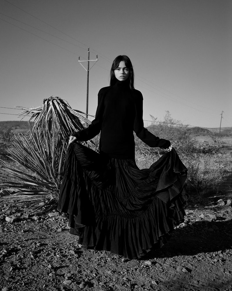 Monochrome portrait of a woman in a black dress standing in a desert landscape with a power pole and sparse vegetation under a clear sky.