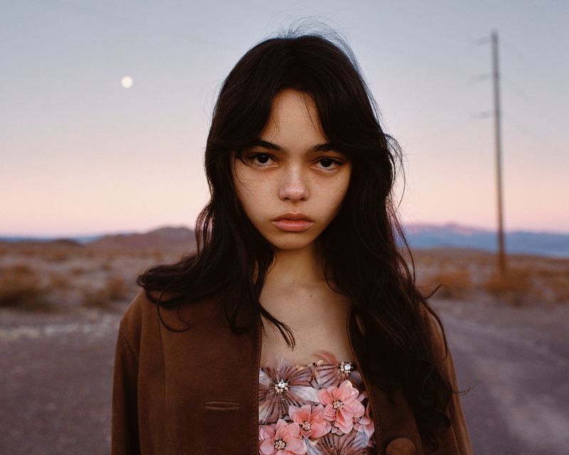 Portrait of a young woman in a floral top and brown jacket posing in a desert landscape under a soft sky with a distant moon.