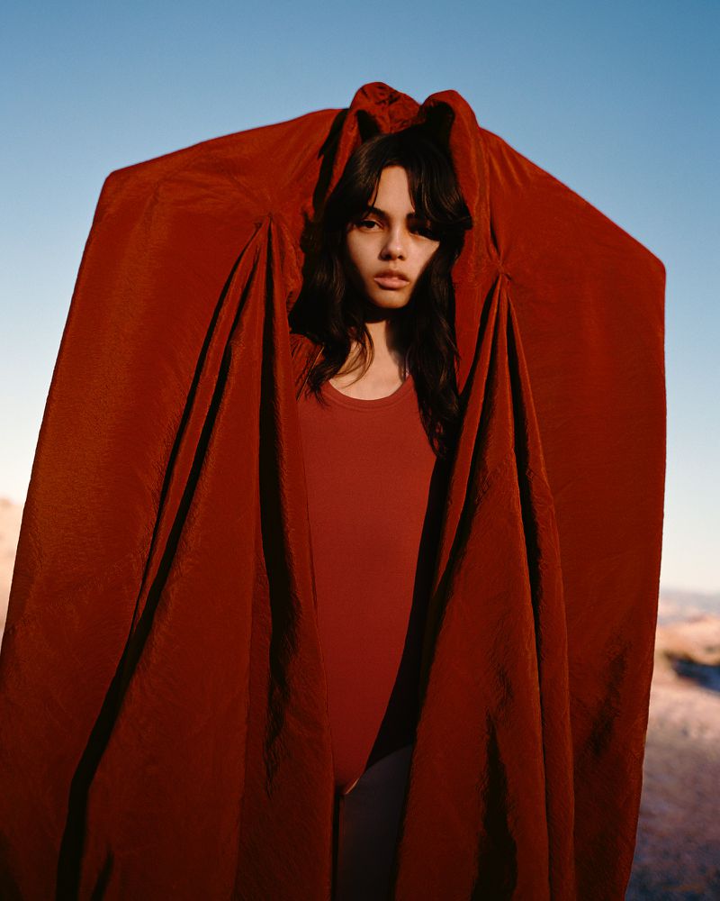 Young woman with dark hair stands outdoors, partially covered by draped rust-colored fabric against a clear blue sky backdrop.