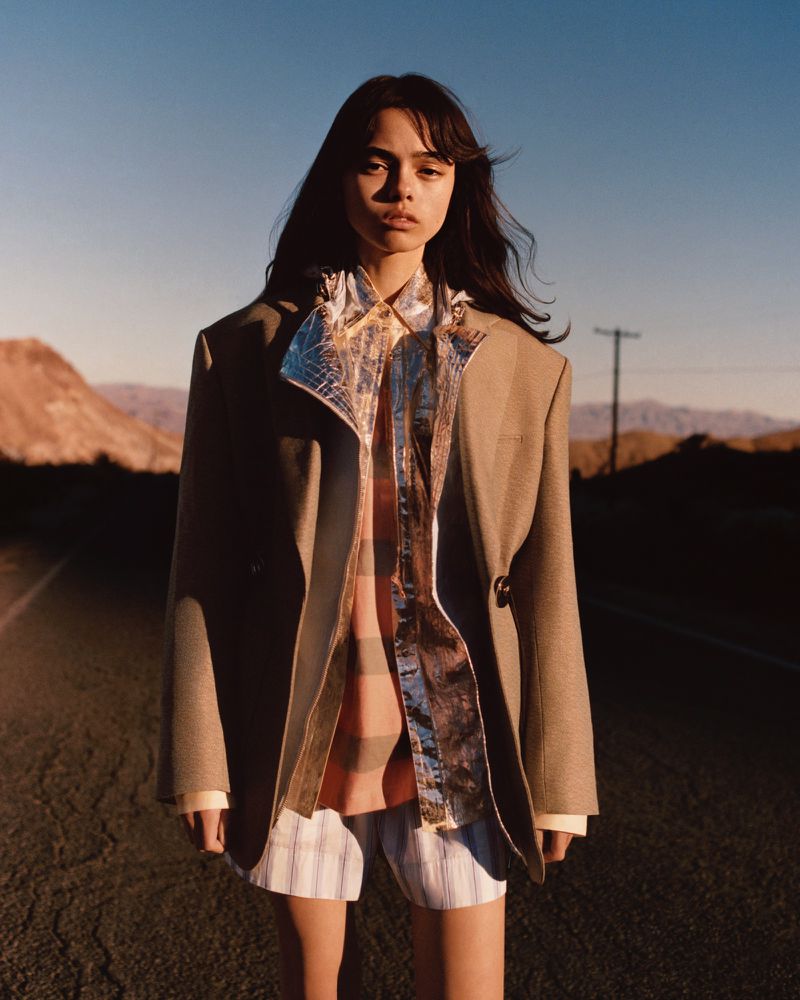 A woman in an oversized blazer and striped shorts standing on a desert road, gazing directly at the camera under a clear blue sky.