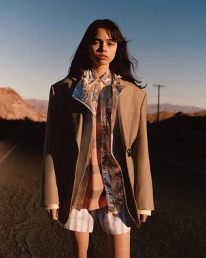 Model standing on a paved road outdoors at sunset wearing a beige blazer over a patterned shirt and shorts.
