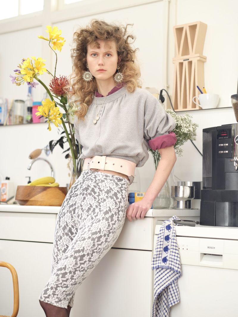 Model Julie Hoomans wearing a grey sweatshirt, wide pink belt, and patterned lace pants, leaning against a white kitchen counter.