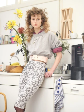Model Julie Hoomans wearing a grey sweatshirt, wide pink belt, and patterned lace pants, leaning against a white kitchen counter.