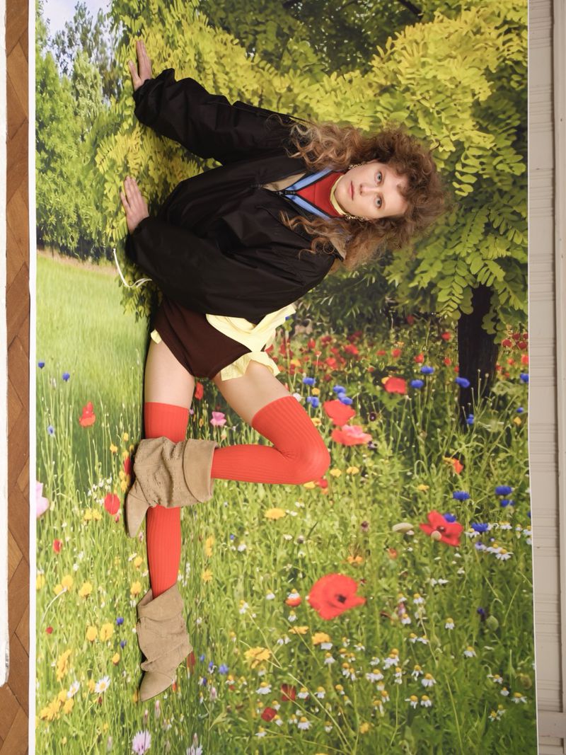 Model Julie Hoomans wearing a black jacket, red tights, and slouchy beige boots posing against a scenic wildflower backdrop.