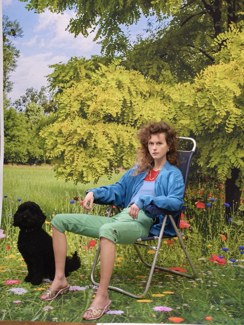 Model Julie Hoomans in a blue jacket and green cropped pants seated in a folding chair next to a black dog in front of a meadow backdrop.