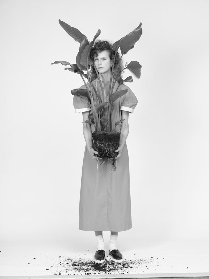 Woman in vintage dress holding potted plant in studio, standing on dirt wearing socks and loafers in a minimalist black and white scene.
