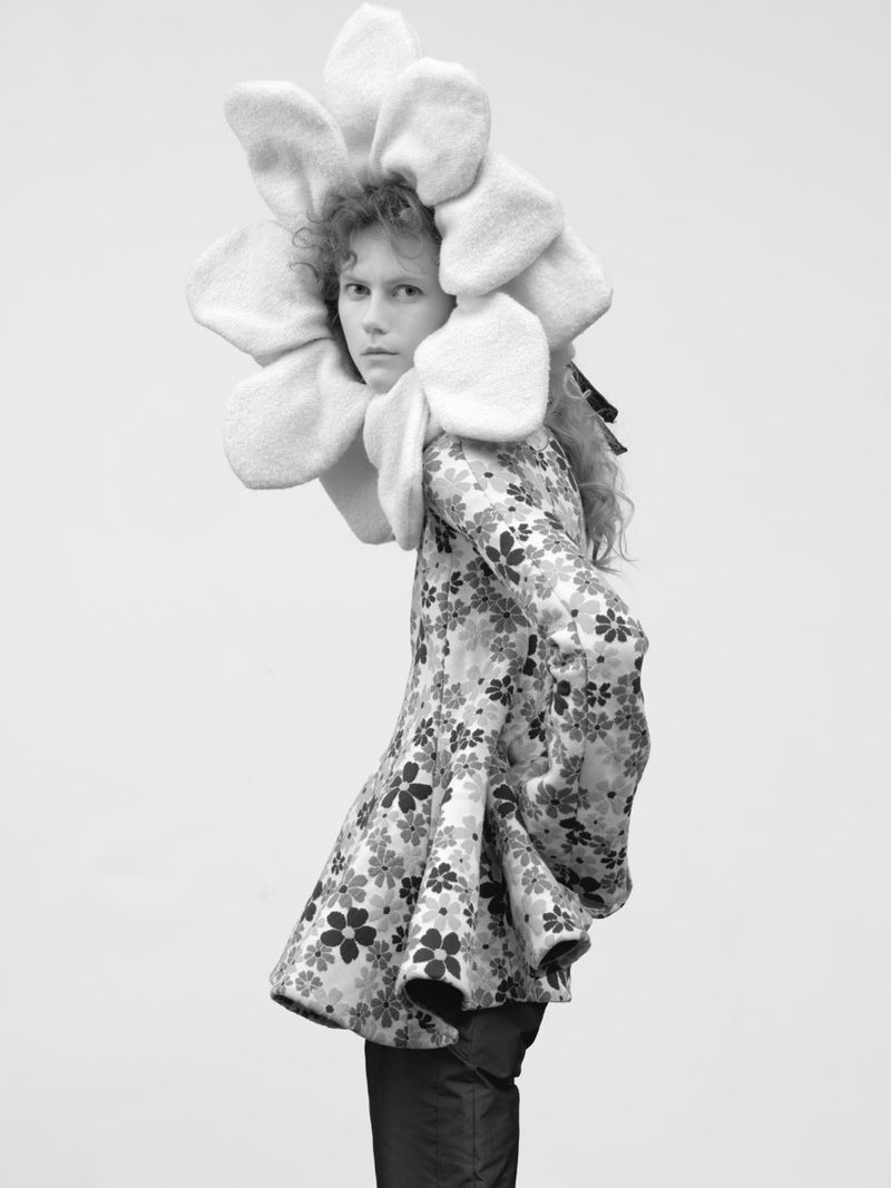 Model Julie Hoomans wearing an oversized plush flower headpiece and floral print jacket against a plain white background.