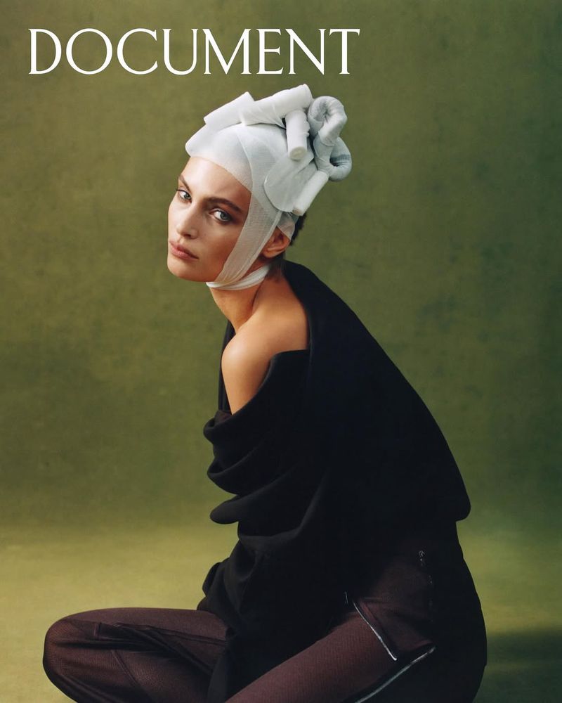 Fashion editorial portrait of a model wearing a headpiece, sitting against an olive green background in a studio setting.