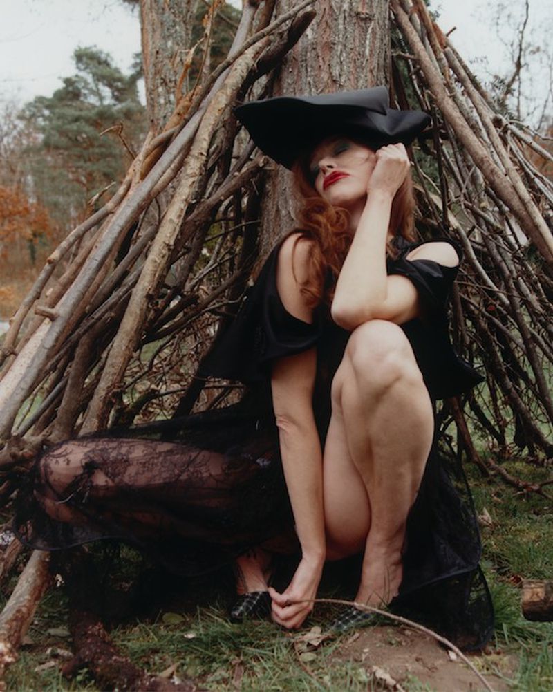 Paz de la Huerta crouching in an all-black dress and sculptural hat beneath a rustic branch shelter in the woods.