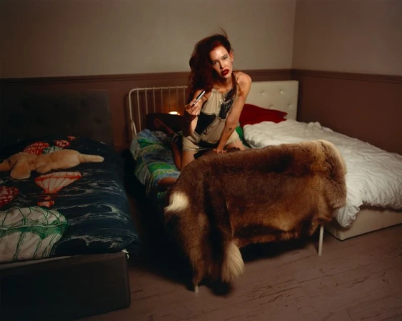 Paz de la Huerta poses on a bed draped in a fur throw wearing a patchwork slip dress in a bedroom setting.
