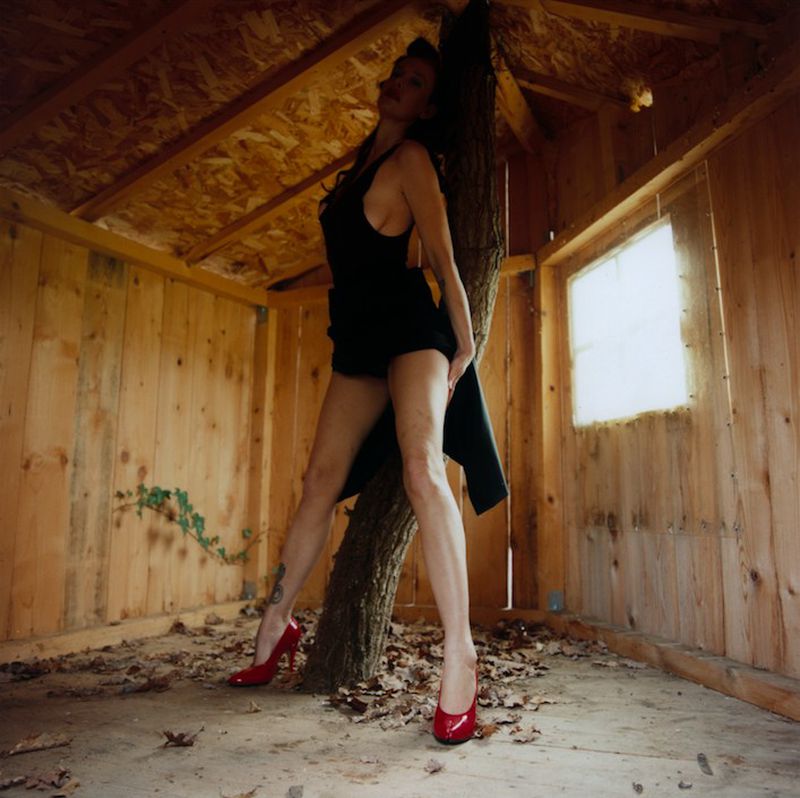 Paz de la Huerta posing in a black bodysuit and red high heels inside a rustic wooden cabin against an indoor tree trunk.