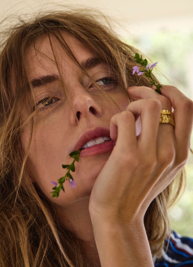 Close-up portrait of a woman with tousled brown hair biting a stem of purple flowers, with a textured gold ring on her finger.