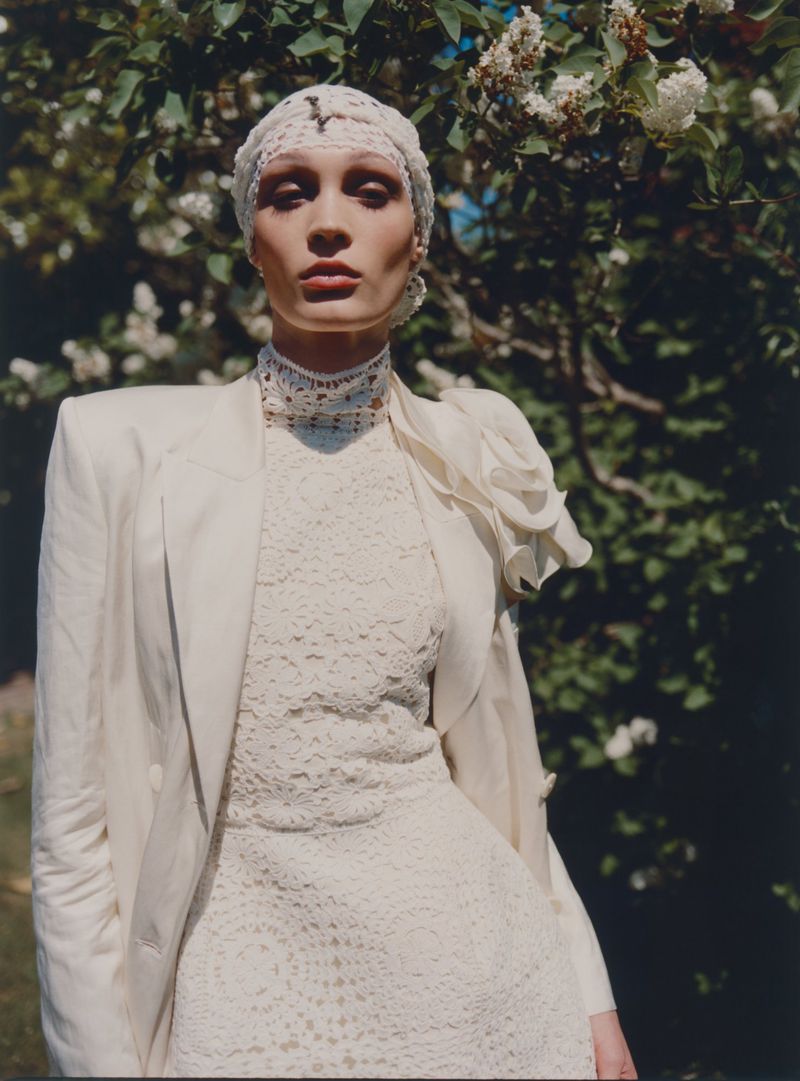 Elegant woman wearing a white lace dress and jacket in a sunlit outdoor setting with a lilac bush in the background.