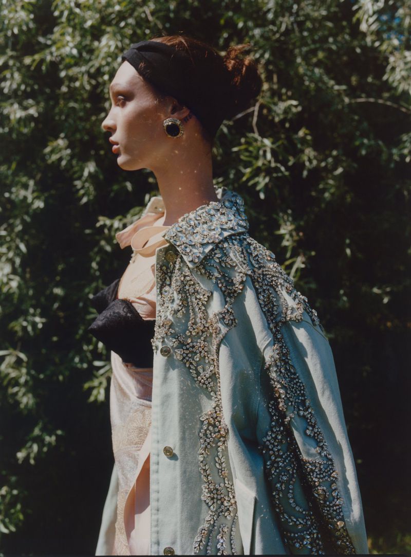 Profile shot of a fashion model with an embellished denim jacket, posing outdoors against a green leafy background in natural light.