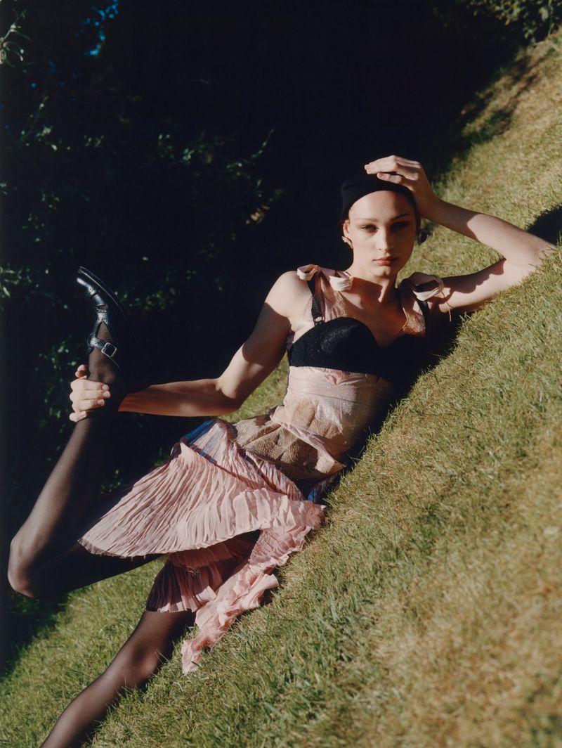 Woman in black top and pink dress lying on grass, leg raised, wearing black Mary Jane shoes. Fashion editorial shot outdoors.