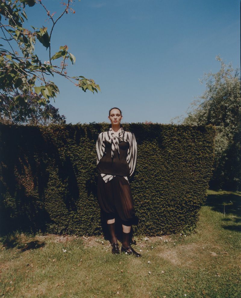 Adult female fashion model poses in a black dress and striped top against a green hedge and blue sky in a natural outdoor setting.