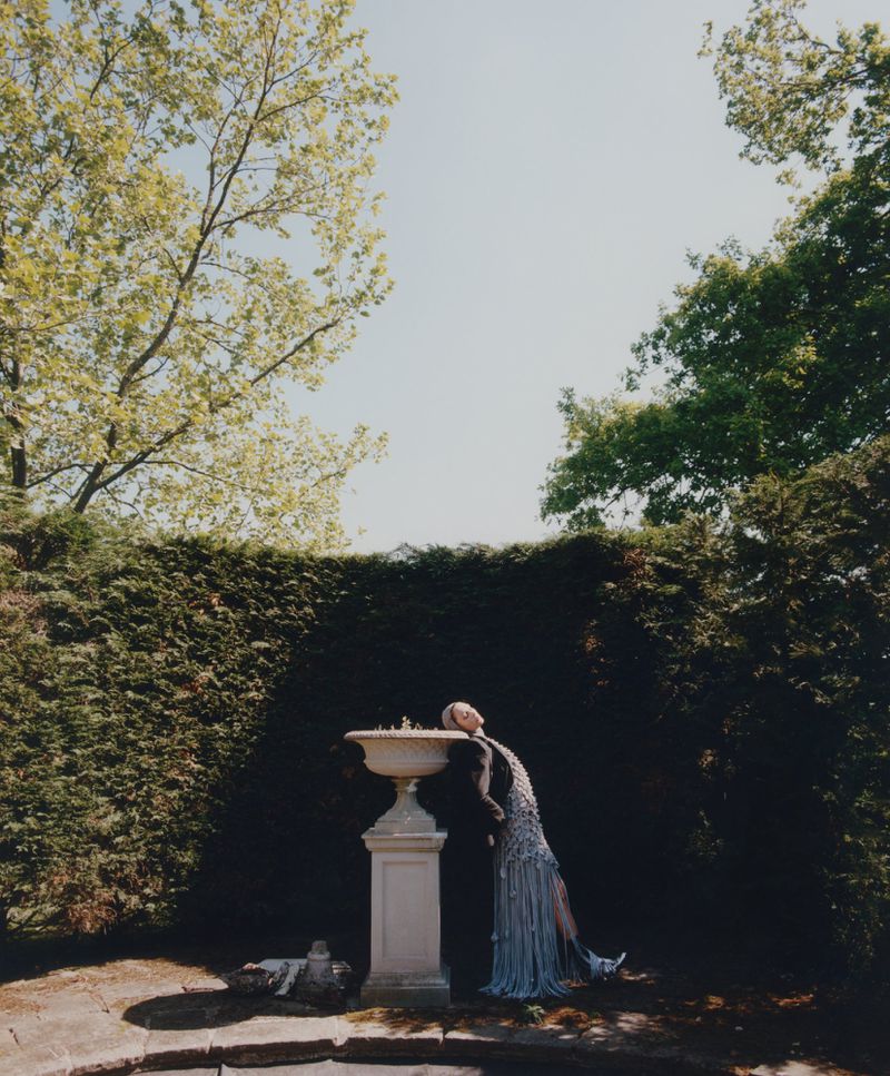 Woman in ornate dress leans head on marble fountain in lush green garden with trees and sky in soft, bright outdoor lighting.