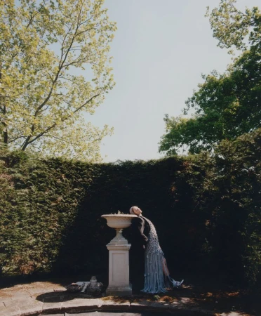 Woman in ornate dress leans head on marble fountain in lush green garden with trees and sky in soft, bright outdoor lighting.