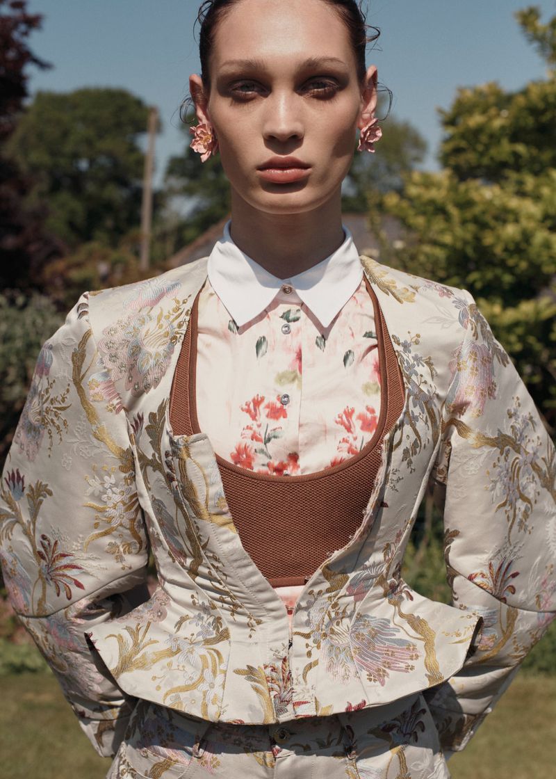 Portrait of a woman in a floral jacket standing in a lush green outdoor garden setting with bright sunlight, wearing floral earrings.