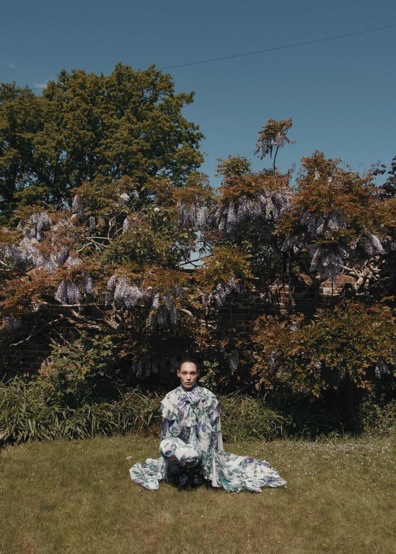 Young woman in a floral dress poses kneeling in a grassy garden surrounded by trees with purple flowers on a sunny day.