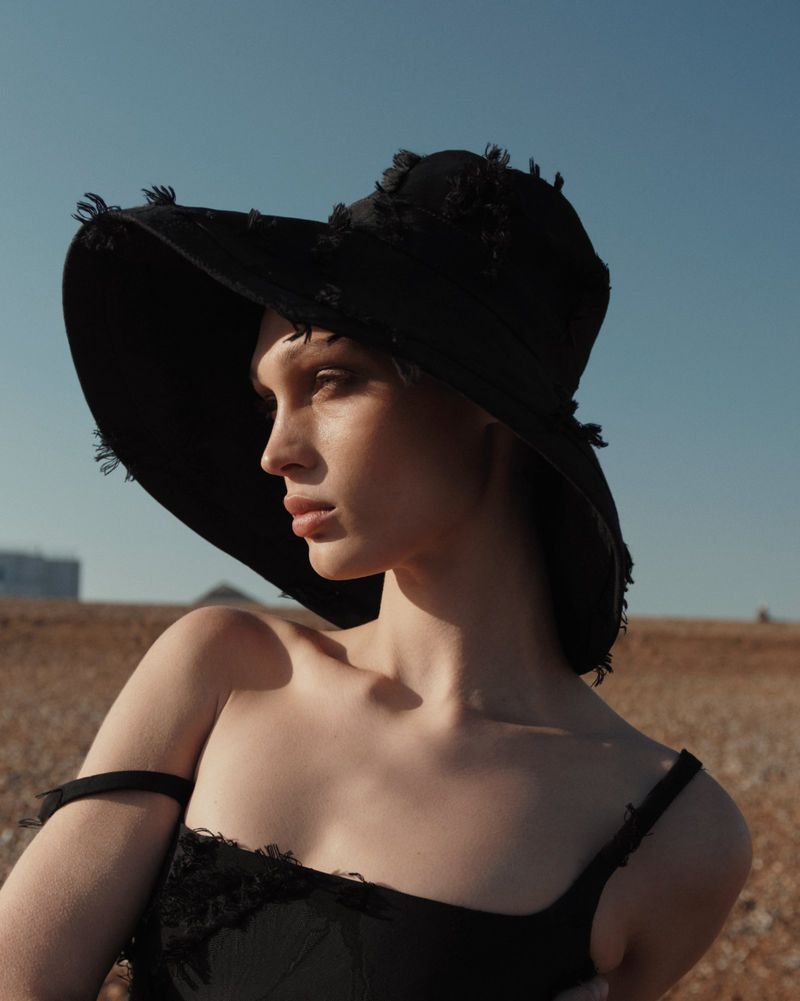 Woman in black hat and dress poses in a field under a blue sky, highlighting fashion, beauty, and outdoor aesthetics.