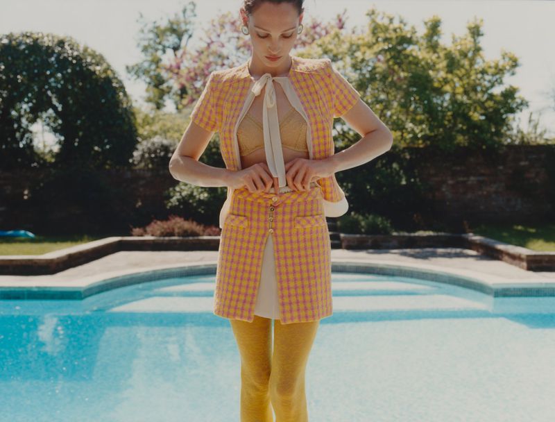 Young adult female poses in a yellow plaid skirt suit by a turquoise swimming pool in a yard with trees and sunlight. 