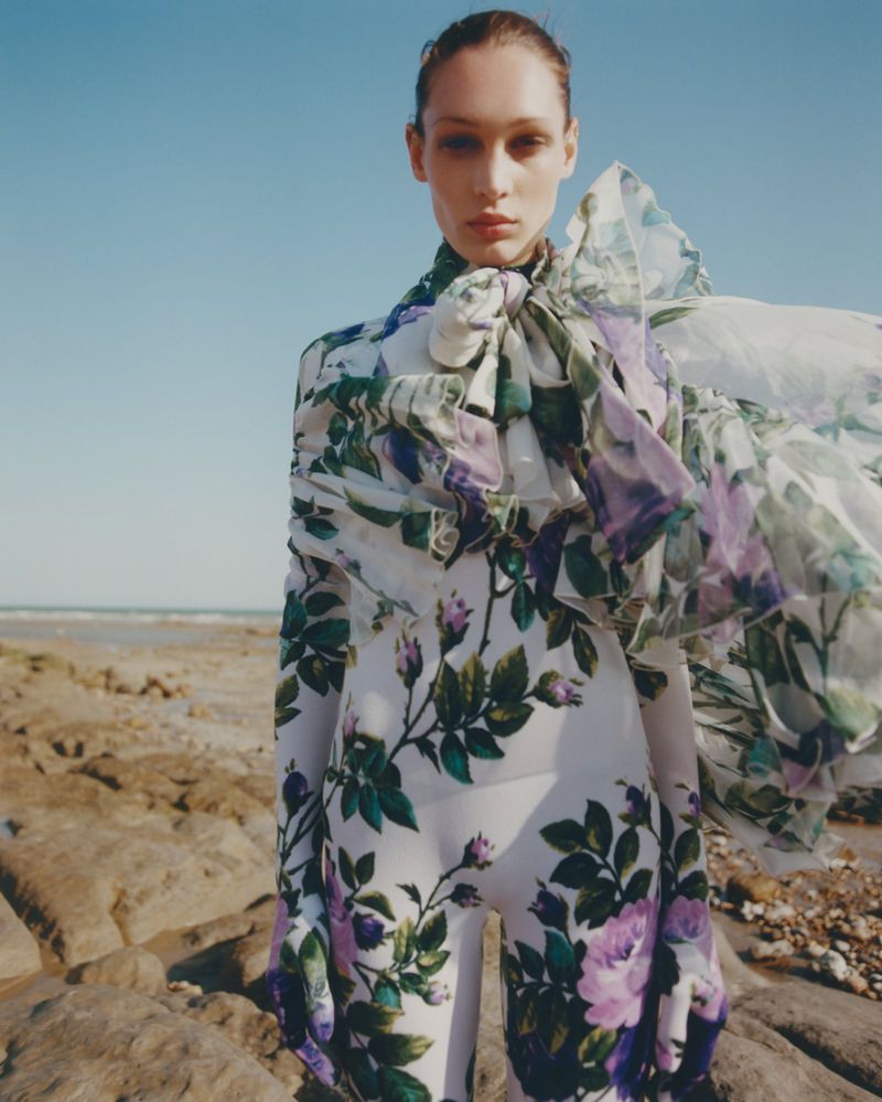 A woman in a full floral-patterned outfit stands on a rocky beach with the ocean and sky in the background, bathed in sunlight.