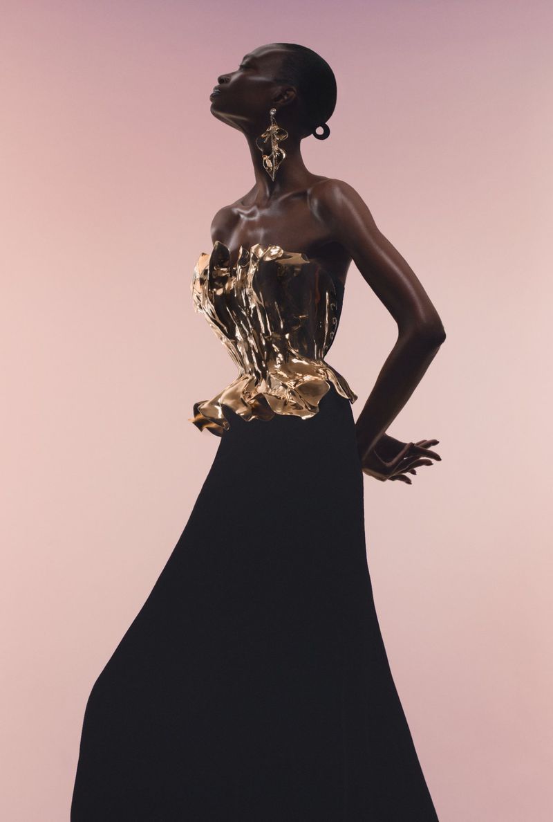 Debra Shaw wearing a sculptural gold metallic corset top and long black skirt against a soft pink studio backdrop.