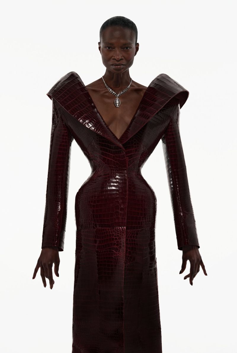 Model Debra Shaw wearing a sculptural burgundy crocodile embossed coat with exaggerated shoulders against a white background.
