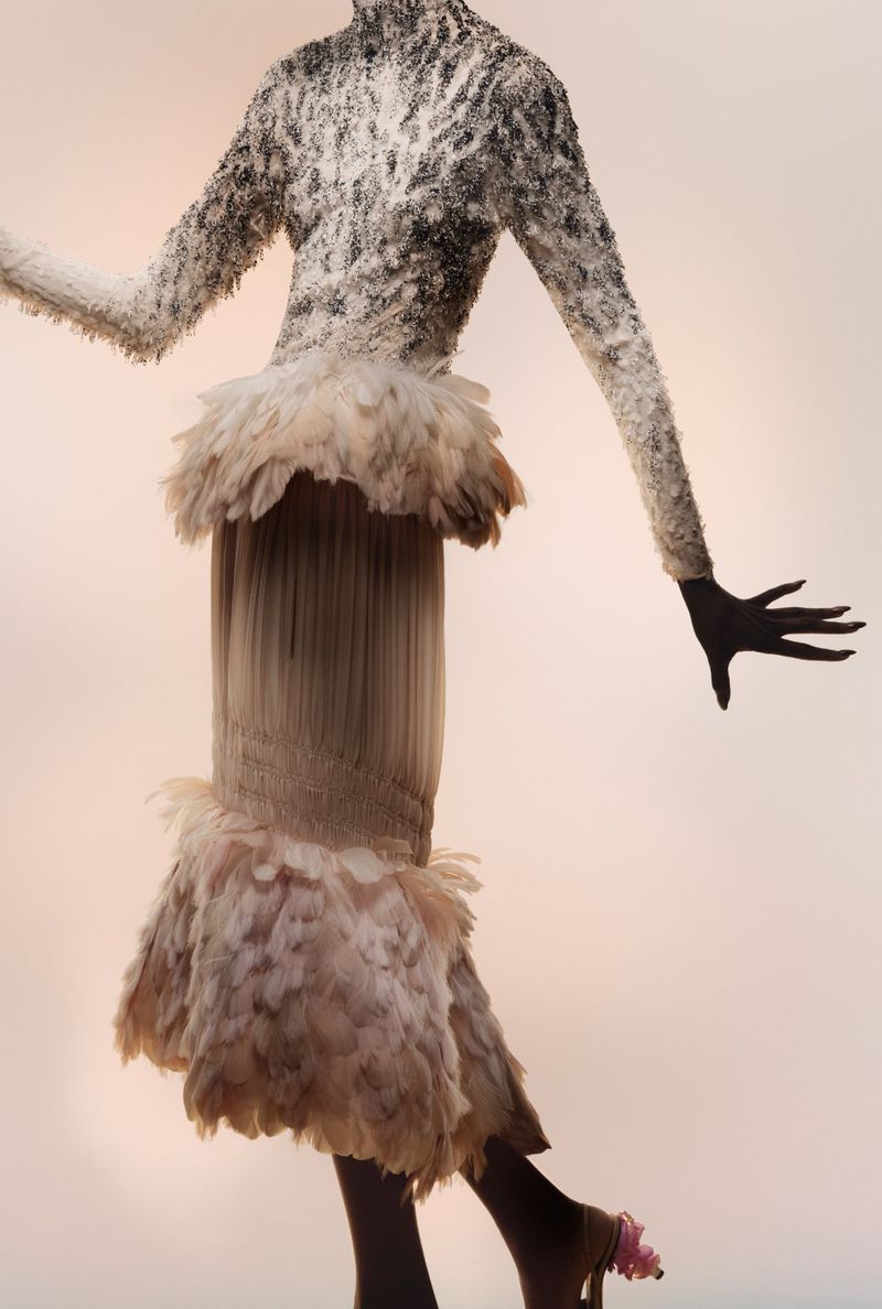 Model Debra Shaw poses in an intricate textured feather dress against a soft backlit studio backdrop.