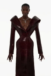 Model Debra Shaw wearing a sculptural burgundy crocodile embossed coat with exaggerated shoulders against a white background.