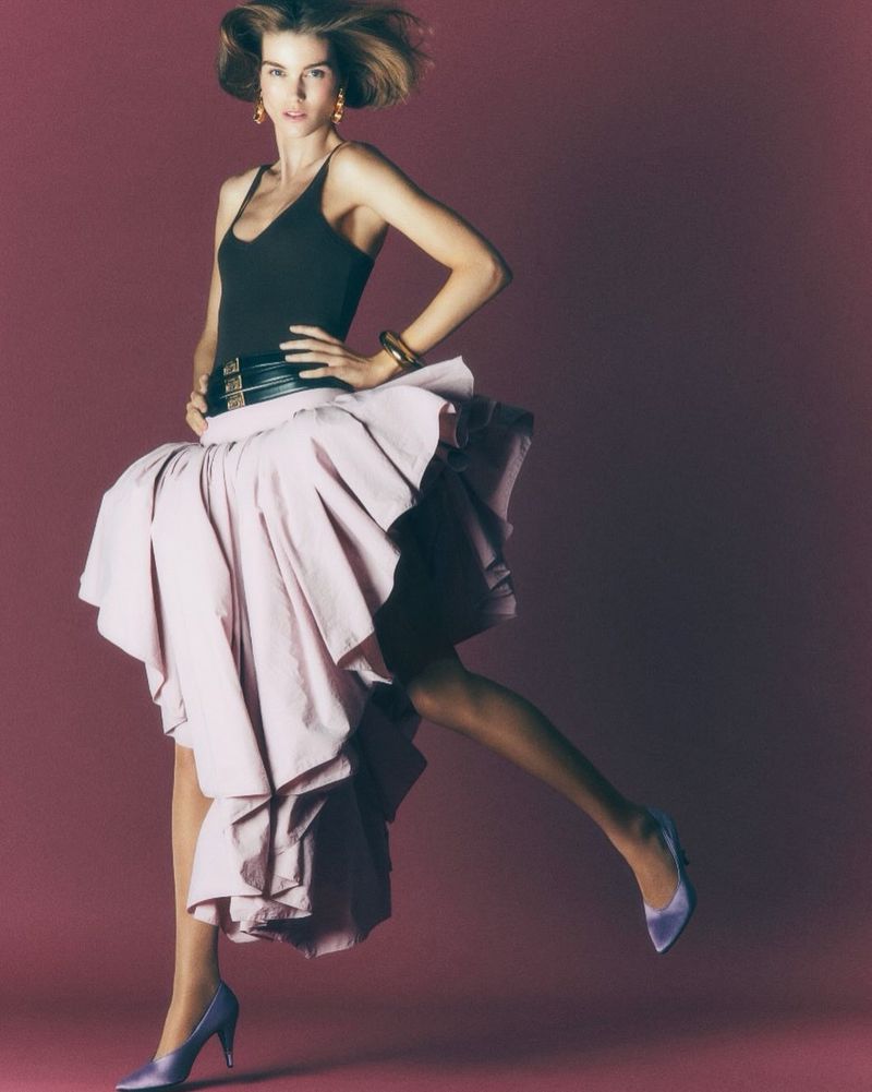 Model Luna Bijl poses mid-motion in a black tank top, voluminous pale pink ruffle skirt, and satin heels against a burgundy backdrop.
