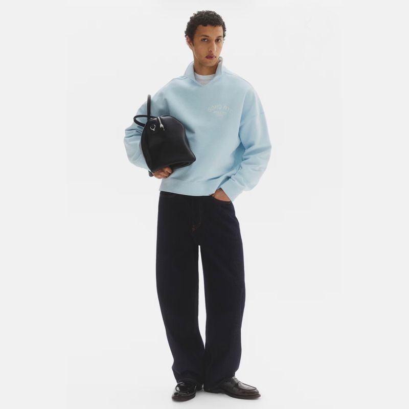 Model Anass Bouazzaoui wearing a light blue sweatshirt and wide-leg dark pants holding a black bag against a white studio background.